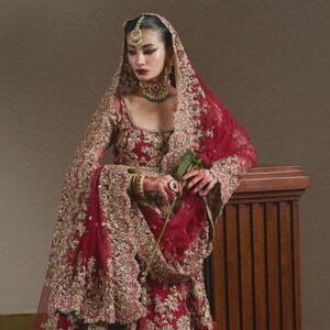 May include: A crimson bridal gown with detailed gold embroidery. The gown has a long-sleeved top, a full skirt, and a matching veil. The model is wearing a green necklace and holding a rose. The gown is a traditional style.