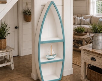 May include: A white wooden bookshelf shaped like a boat with a teal trim. The bookshelf features three open shelves, with a small decorative model sailboat placed on the middle shelf. The structure is tall, narrow, and stands upright on the floor.