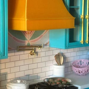 Puede incluir: Una escena de cocina en miniatura con una campana extractora de color amarillo brillante, armarios turquesa y una placa para salpicaduras de azulejos blancos. Una fuente blanca para hornear, una estufa de gas y una sartén con comida están en la encimera. También se ve una cesta rosa y un soporte blanco para utensilios.