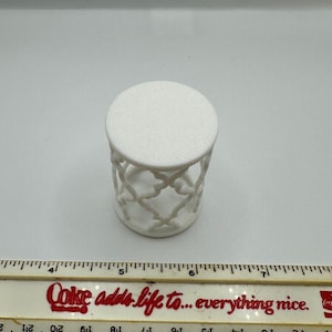 Pu&ograve; includere: Tavolino decorativo bianco con piano rotondo e design traforato sui lati. Il tavolino &egrave; alto circa 7.6 cm. Un righello con la scritta "Coke adds life to... everything nice" &egrave; in primo piano.