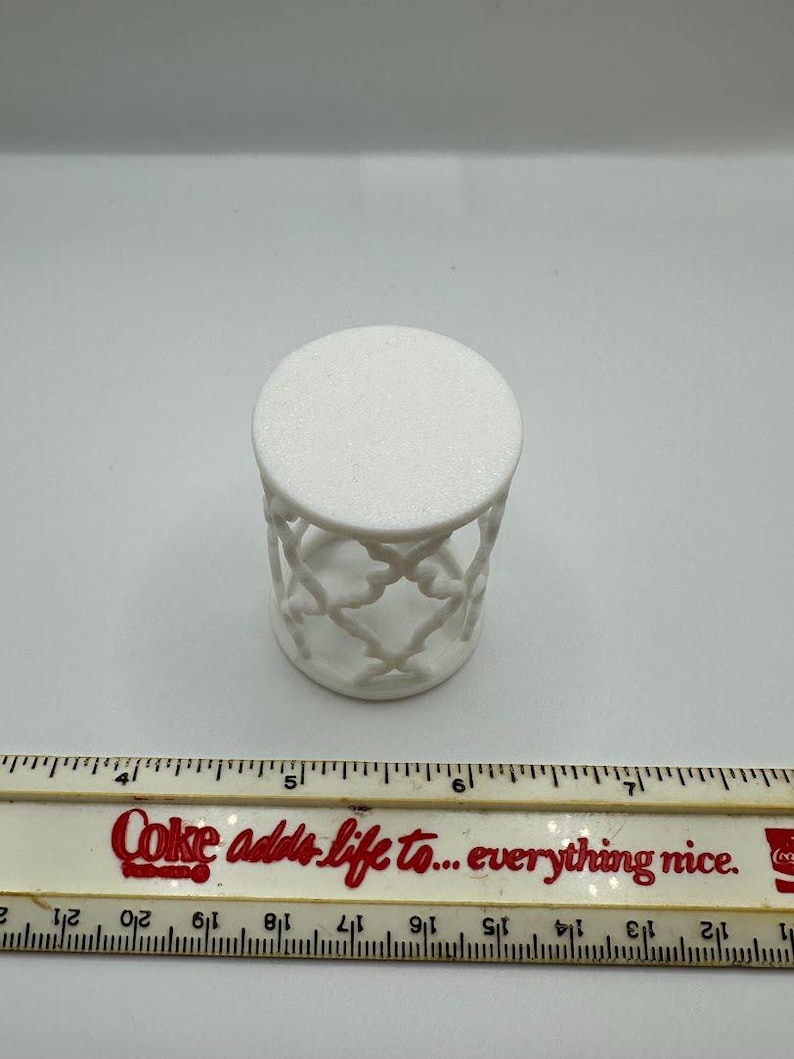Pu&ograve; includere: Tavolino decorativo bianco con piano rotondo e design traforato sui lati. Il tavolino &egrave; alto circa 7.6 cm. Un righello con la scritta "Coke adds life to... everything nice" &egrave; in primo piano.