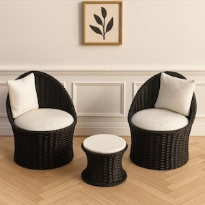 May include: Two black wicker chairs with white cushions and pillows, and a matching side table. A framed artwork with a leaf design hangs on the wall above the chairs. The furniture is arranged on a light wood floor.