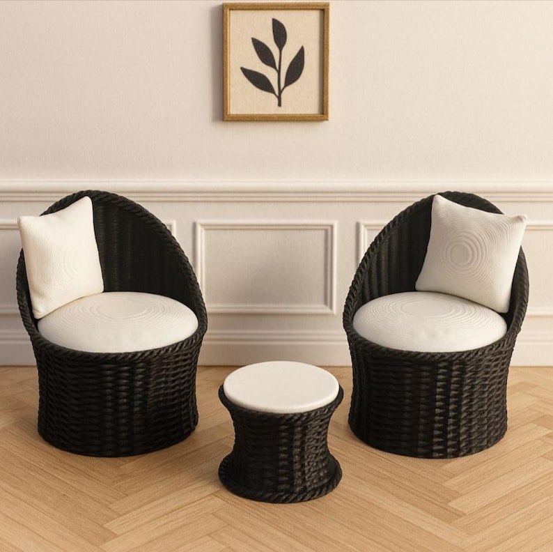 May include: Two black wicker chairs with white cushions and pillows, and a matching side table. A framed artwork with a leaf design hangs on the wall above the chairs. The furniture is arranged on a light wood floor.