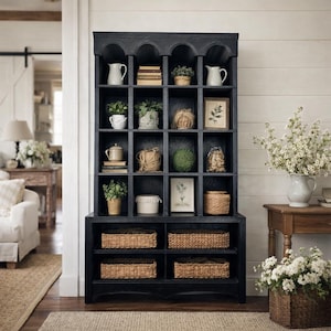 May include: A black cabinet with multiple shelves, featuring an arched top, and filled with various decorative items. The shelves display plants, books, pitchers, and framed art. Woven baskets are in the lower section. The cabinet is in a bright room.