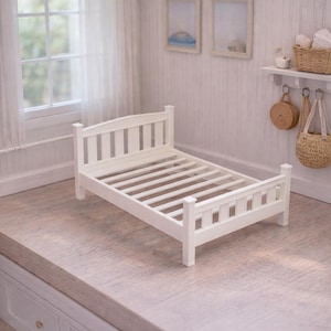 May include: A white wooden doll's bed frame with a slatted base. The bed is in a miniature bedroom setting with a window, curtains, and decorative wall art. The room has a neutral colour palette.