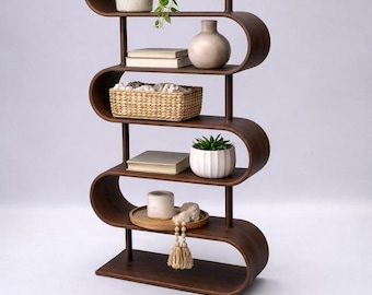 May include: A dark brown, modern, curved bookshelf with multiple shelves. The shelves display various decorative items, including plants in pots, books, a vase, and a woven basket. The design is minimalist and stylish.