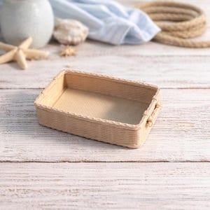 May include: A rectangular woven basket with a light beige color. The basket has a textured surface and small handles on each side. The background includes a vase of white flowers, seashells, and rope, suggesting a nautical theme.