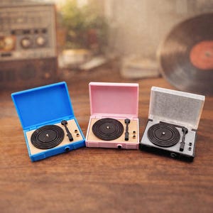 May include: Three miniature record players in blue, pink, and grey, each with a black record and tone arm. The record players are open, revealing the turntable. The background includes a vintage radio and a vinyl record.
