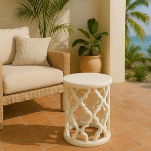 May include: A white, round side table with a decorative, openwork design. The table is made of a solid material and has a smooth surface. The table is placed on a terracotta tile patio, next to a wicker chair and potted plants.