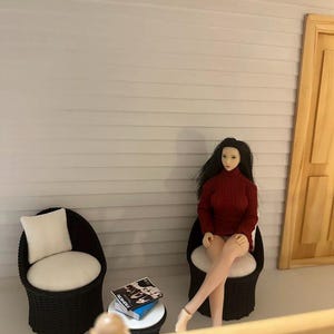 May include: Miniature scene featuring a doll in a red turtleneck sweater seated in a wicker chair. A small white table holds books with the word "FASHION" visible. Another wicker chair with a white cushion is also present. The scene is set against a light gray wall.