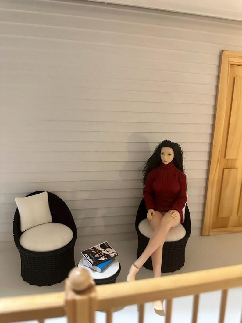 May include: Miniature scene featuring a doll in a red turtleneck sweater seated in a wicker chair. A small white table holds books with the word "FASHION" visible. Another wicker chair with a white cushion is also present. The scene is set against a light gray wall.