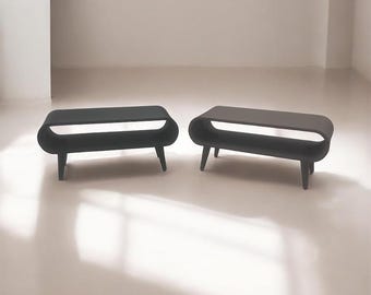 May include: Two black, modern-style coffee tables. Each table has a rounded rectangular top with a lower shelf and four short, tapered legs. The tables are made of a solid, dark material.