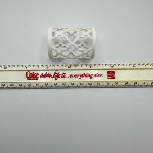 Pu&ograve; includere: Un oggetto cilindrico bianco con un design traforato decorativo. L'oggetto &egrave; lungo circa 5 cm e si trova su una superficie bianca accanto a un righello. Il righello riporta il testo "Coke adds life to... everything nice."