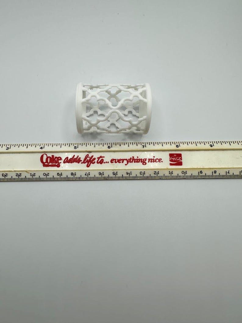 Pu&ograve; includere: Un oggetto cilindrico bianco con un design traforato decorativo. L'oggetto &egrave; lungo circa 5 cm e si trova su una superficie bianca accanto a un righello. Il righello riporta il testo "Coke adds life to... everything nice."