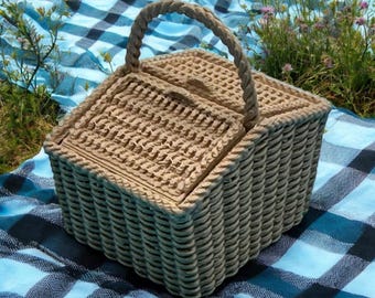 Op de afbeelding: Een beige geweven picknickmand met een bijpassend deksel en een touw handvat. De mand staat op een blauw-zwarte geruite deken, met gras en wilde bloemen op de achtergrond.