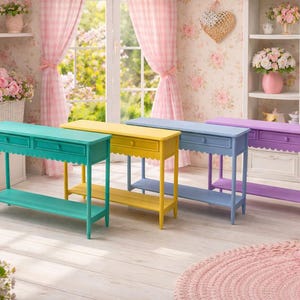 Op de afbeelding: Vier kleurrijke consoletafels in teal, geel, blauw en paars. Elke tafel heeft een lade, een geschulpte rand en een onderste plank. De tafels staan in een lichte kamer met bloemen.