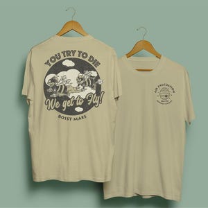 May include: Two beige t-shirts hanging on wooden hangers. One shirt features a circular graphic with bees and the text "YOU TRY TO DIE, WE GET TO FLY!" and "801ST MAES." The other shirt has a bee-themed logo with the text "AIR EVACUATION" and "HUMAN FLYERS."