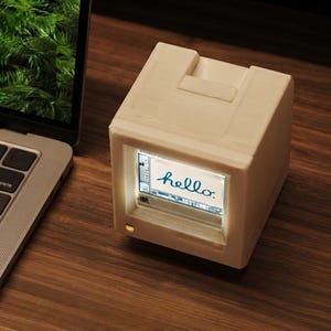 May include: A white, cube-shaped electronic device with a small, recessed screen displaying the word hello in a blue script font. The device features a matte finish, a small indicator light at the bottom, and a top-mounted slot, resembling a miniature computer.