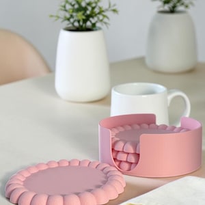 Può includere: Un set di sottobicchieri rotondi rosa con un design a bordo smerlato. I sottobicchieri sono realizzati in materiale opaco e sono riposti in un supporto cilindrico rosa abbinato con un'apertura laterale. Un sottobicchiere è appoggiato in piano.