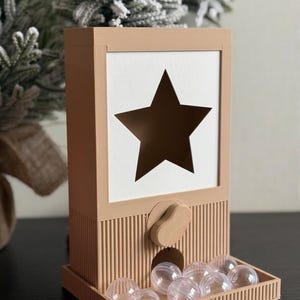 Pode incluir: Um dispensador retangular bege com um painel frontal branco que apresenta um grande recorte central em forma de estrela. A base tem uma textura estriada, um botão giratório manual e uma bandeja contendo vários pequenos recipientes esféricos de plástico transparente.
