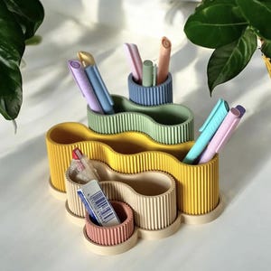 Op de afbeelding: Een bureau-organizer met meerdere niveaus en een geribbelde textuur, bestaande uit onderling verbonden golvende vakken in geel, saliegroen, blauw, beige en roze. De organizer bevat diverse pennen en een witte gum met een barcode.