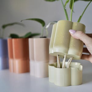 Op de afbeelding: Een lichtgele zelfbewaterende plantenpot met een geribbelde, geschulpte buitenkant en een bijpassend basisreservoir verbonden door twee witte katoenen lonten. De pot bevat een groene plant, met andere soortgelijke potten in paars, oranje en beige zichtbaar.