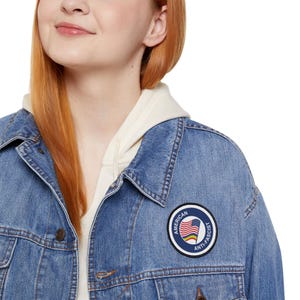 Può includere: Giacca di jeans blu chiaro con una toppa rotonda con la scritta "AMERICAN ANTI-FASCIST" attorno a un disegno a bandiera arcobaleno. La giacca è indossata sopra una felpa con cappuccio color crema.