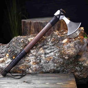 May include: A hand axe featuring a polished silver metal blade with a curved edge, a dark brown wooden handle, and a black leather-wrapped grip. The head of the axe is secured with a black cross-patterned binding near the top of the handle.