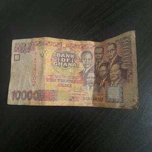 May include: A Ghanaian ten thousand Cedis banknote. The bill features portraits of several men and the text "BANK OF GHANA" and "TEN THOUSAND CEDIS". The bill is primarily yellow, red, and brown.