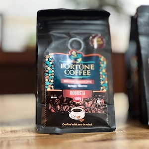 Puede incluir: Bolsa negra de granos de café Robusta Fortune Coffee. La bolsa tiene una etiqueta turquesa y roja con el texto "Fortune Coffee", "Wholesome Ghanaian Coffee" y "Naturally Processed". La bolsa contiene 250g de café.