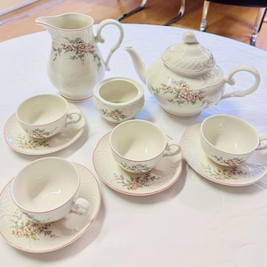 May include: A white porcelain tea set with floral accents. The set includes a teapot, creamer, sugar bowl, and six teacups with saucers. Each piece features a delicate pink and green floral pattern and a red trim.