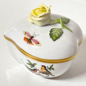 May include: A heart-shaped, white porcelain trinket box with a yellow rose on the lid. The box features a gold trim and is decorated with a butterfly and birds. The rose stem and leaves are green.