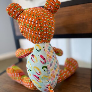 May include: A handmade fabric bear doll with orange patterned fabric on the head, ears, and limbs. The body features a light blue fabric with colorful ring designs and a section with a geometric pattern. The bear is sitting on a wooden surface.