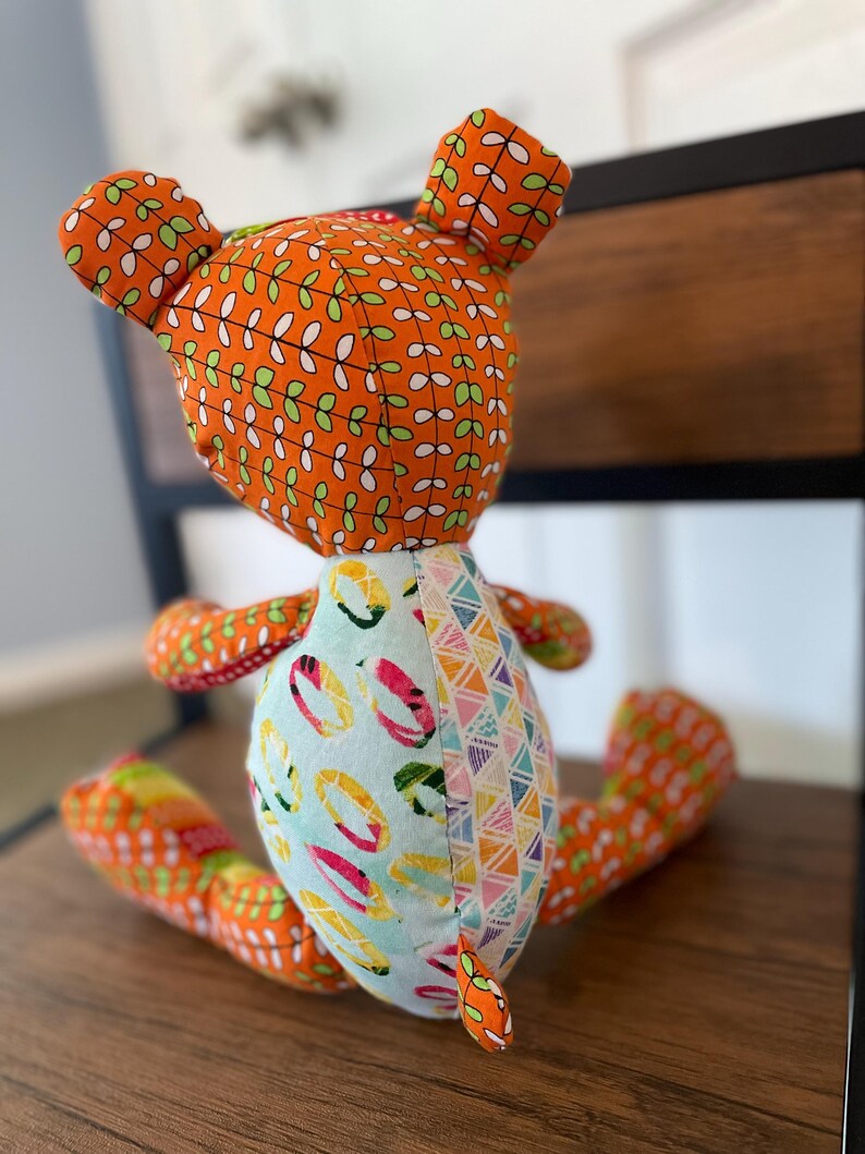May include: A handmade fabric bear doll with orange patterned fabric on the head, ears, and limbs. The body features a light blue fabric with colorful ring designs and a section with a geometric pattern. The bear is sitting on a wooden surface.