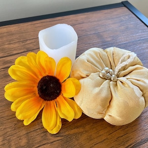 May include: A white cylindrical candle sleeve, a bright yellow sunflower with a dark brown center, and a round, beige fabric pumpkin ornament featuring a small white loop at the top and a cluster of five small, round, cream-colored faux pearls.