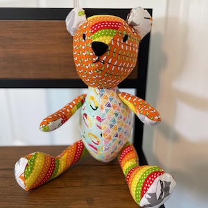 May include: A handmade stuffed bear with a patchwork design. The bear features orange, green, yellow, and red fabrics with leaf and geometric patterns. It has a black nose and small ears. The bear is sitting on a wooden surface.