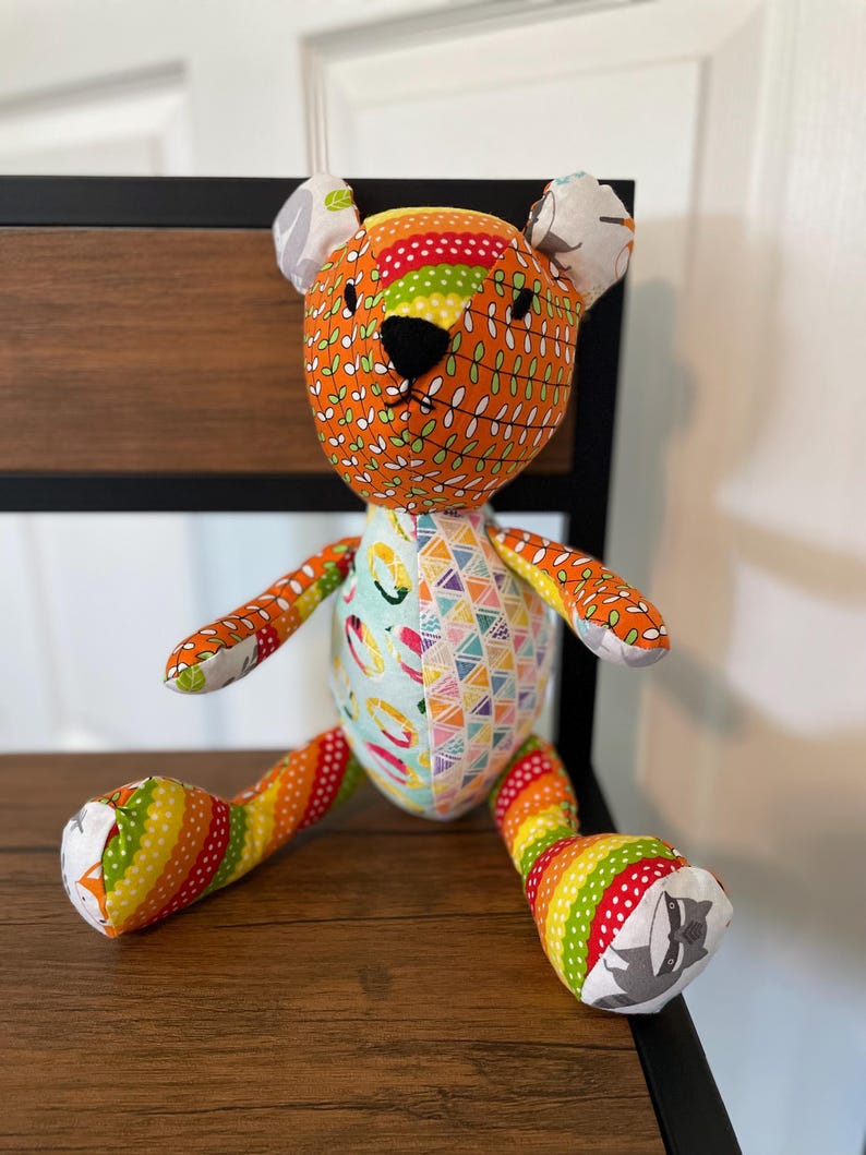 May include: A handmade stuffed bear with a patchwork design. The bear features orange, green, yellow, and red fabrics with leaf and geometric patterns. It has a black nose and small ears. The bear is sitting on a wooden surface.