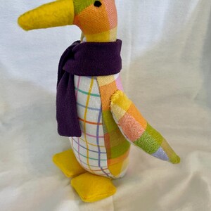 May include: A stuffed penguin toy crafted from various fabric patterns, including a yellow, orange, and green herringbone weave, and a white grid pattern. The penguin features a solid yellow beak and feet, and wears a dark purple fabric scarf.