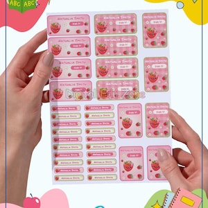 Puede incluir: Una hoja de etiquetas adhesivas rectangulares y cuadradas con un patrón de cuadros vichy rosa, un personaje de fresa de dibujos animados y el texto impreso "NATHALIA SMITH" y "Grade 4". Las etiquetas están dispuestas en varios tamaños.
