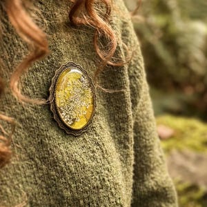 May include: An oval-shaped brooch with a yellow and white floral design set in a bronze-colored frame. The brooch is pinned to a green sweater. The background is a blurred view of a forest.