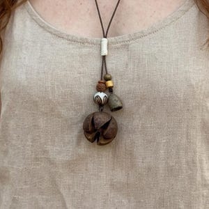 Puede incluir: Un collar largo de cordón marrón con un colgante que presenta un grupo de elementos naturales. El colgante incluye una cápsula de semilla marrón grande y texturizada, pequeñas cuentas de madera y un dije en forma de campana. El collar se usa sobre una blusa de tono neutro.