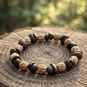 Puede incluir: Un brazalete de cuentas con cuentas de color marrón oscuro y beige alternadas. El brazalete se muestra sobre una superficie de madera con un fondo borroso de bosque. Las cuentas tienen una apariencia texturizada distintiva. El brazalete probablemente está hecho de materiales naturales.
