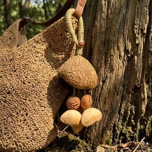 May include: A woven straw shoulder bag with a brown leather strap and a decorative charm. The charm features a large, textured, brown pendant with smaller, light-colored, mushroom-shaped accents. The bag is set against a natural, outdoor backdrop.