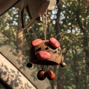 May include: A handmade car charm featuring a pig design. The charm is made from a coconut shell with red and brown accents. It hangs from a brown string, adding a touch of whimsy to any vehicle's interior.