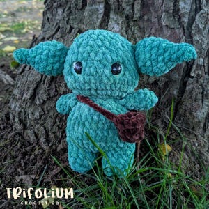 May include: A teal green crocheted plush toy with large ears, black eyes, and a small brown satchel. The toy is made of a textured yarn and is positioned in front of a tree trunk. The logo "Trifolium Crochet Co" is at the bottom.