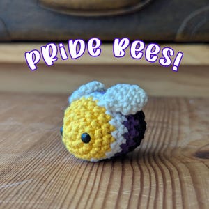 Puede incluir: Una abeja de ganchillo con rayas amarillas, blancas y moradas. La abeja tiene ojos negros y alas blancas. El texto "pride bees!" está encima de la abeja en morado.