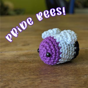 May include: A handmade crochet bee in purple, white, and black, with the text "Pride bees!" in a purple font. The bee has black bead eyes and white wings. The bee is sitting on a wooden surface.