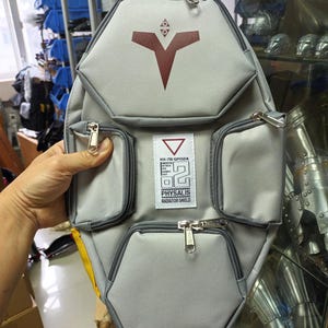 May include: Gray backpack with multiple zippered compartments and a geometric design. Features a red emblem and text that reads "RX-78 GPO2A PHYSALIS RADIATOR SHIELD". The bag has a yellow strap and silver zippers.