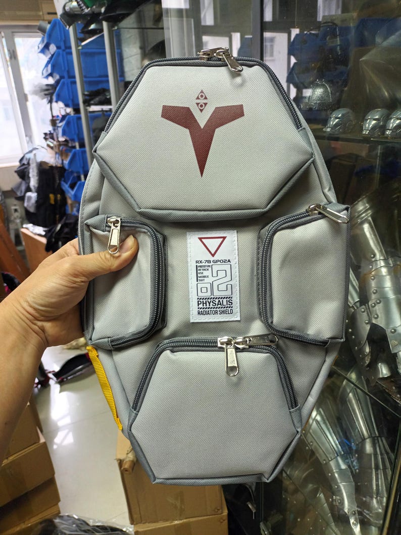 May include: Gray backpack with multiple zippered compartments and a geometric design. Features a red emblem and text that reads "RX-78 GPO2A PHYSALIS RADIATOR SHIELD". The bag has a yellow strap and silver zippers.