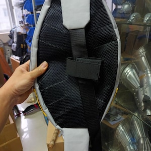 May include: A gray and black sling backpack with a padded back panel and adjustable strap. The backpack features a mesh back for breathability and a gray accent on the top and bottom. The strap has a velcro closure.